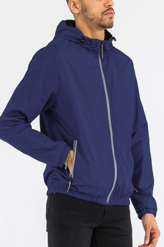 An angled view of a male model wearing the reflective navy blue zip up wind breaker jacket.
