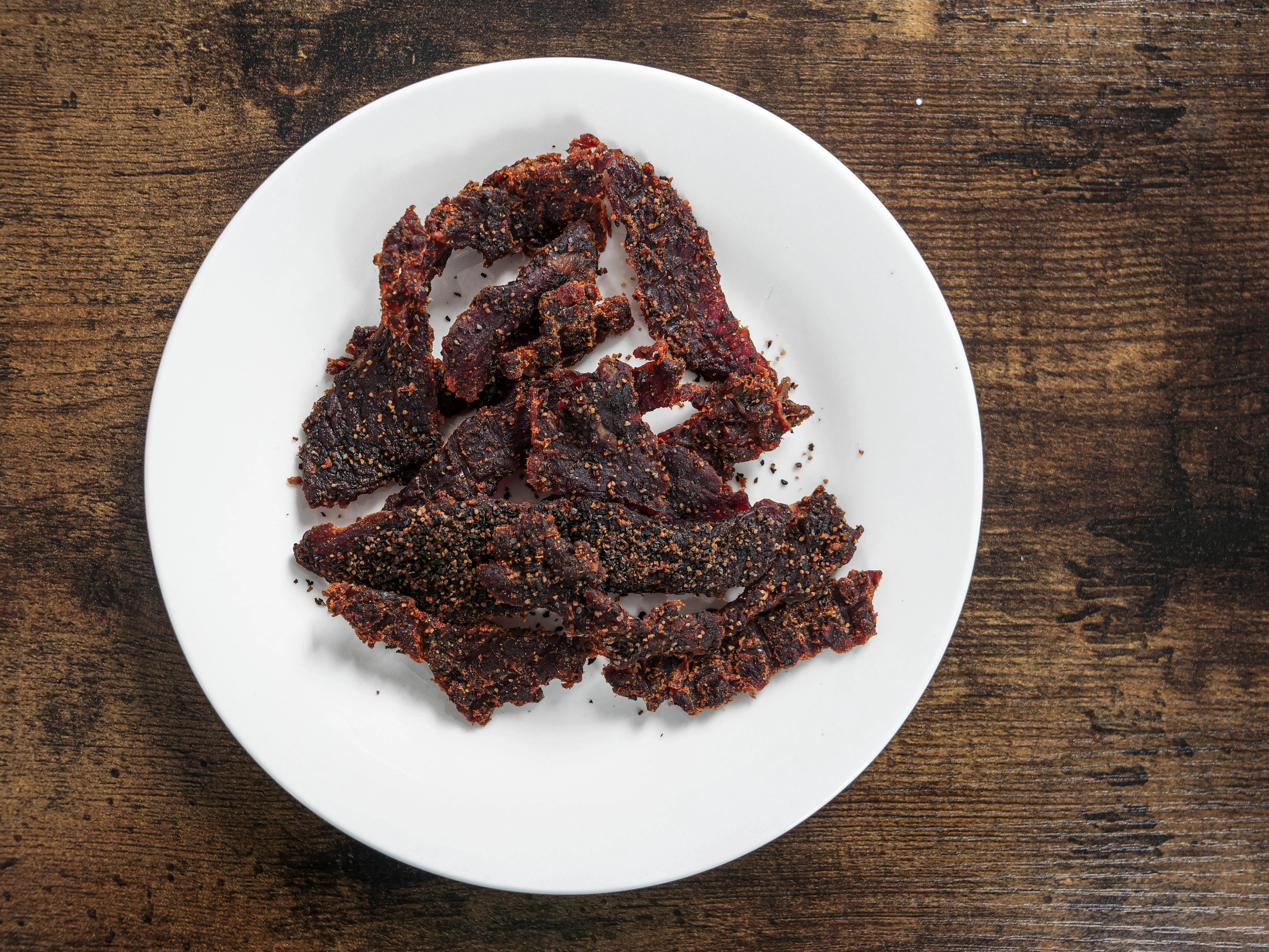 Tong Jerky Black Pepper Sea Salt Beef Jerky displayed on a white plate set upon a brown table. The beef looks well seasons and of a familiar deliciously inviting color. The granules of salt and pepper are distinguishable in this high-quality photograph.