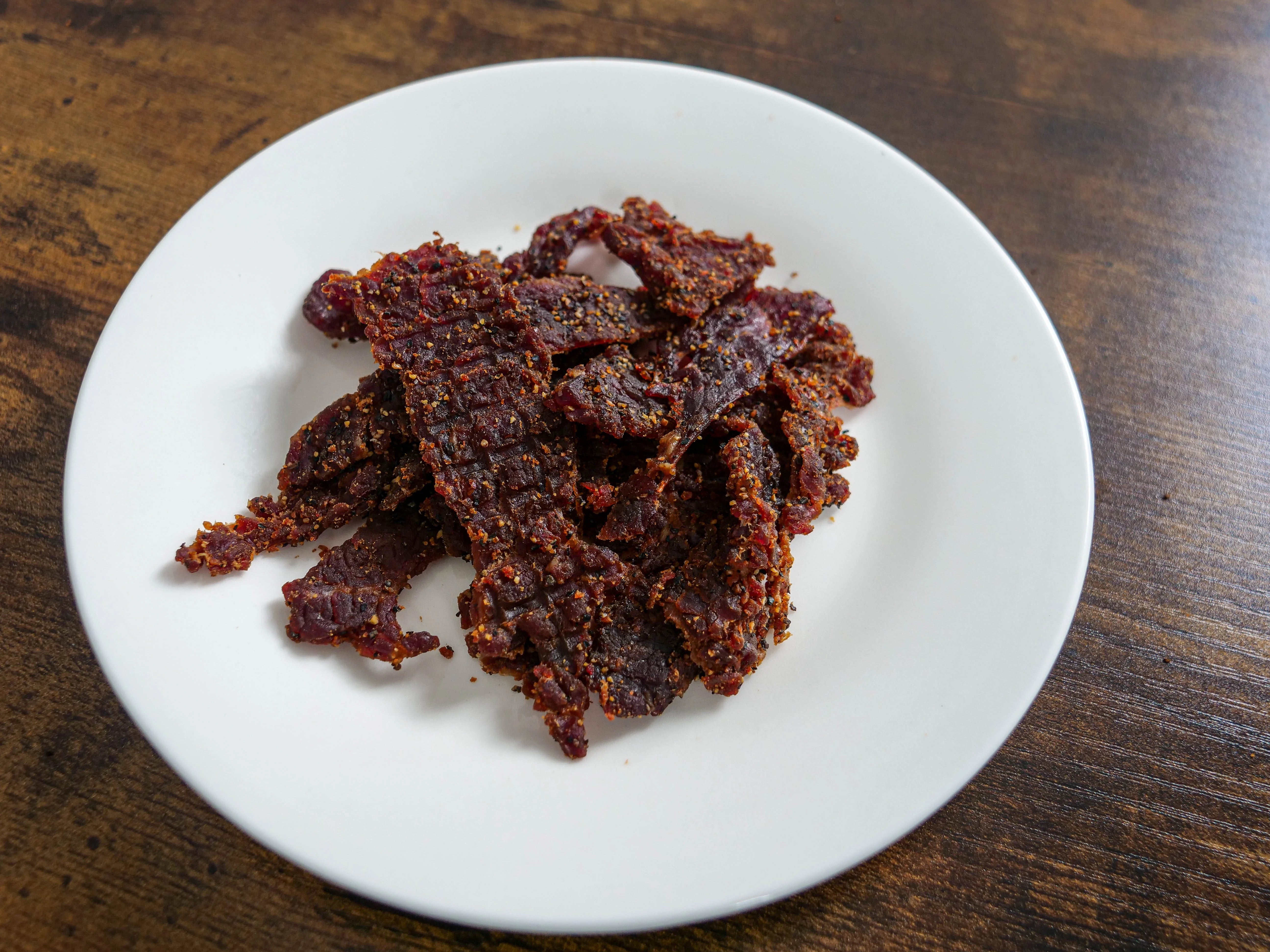 Pieces of soft-textured Tong Jerky Honey Pepper Teriyaki spread out on a white plate set upon a wood table, clearly showing the visible beef jerky, dark pepper seasoning, and glaze.