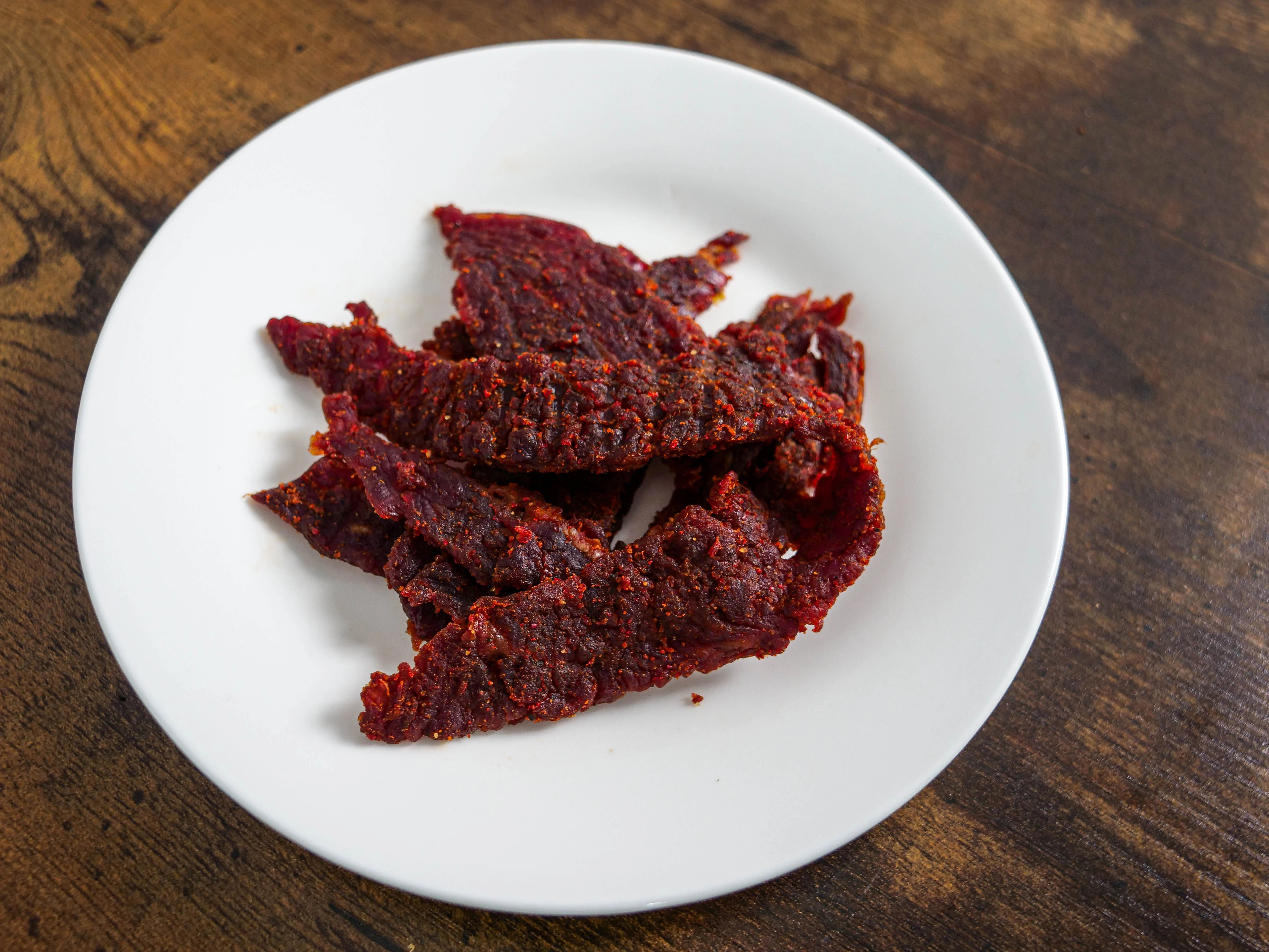 Tong Jerky Spicy Sriracha flavor displayed on a white plate set upon a wooden table. The Beef looks to have a red color due to the sriracha and red chili peppers seasonings and appears seasoned well with grains of salt and pepper distinguishable in the high-quality photograph.