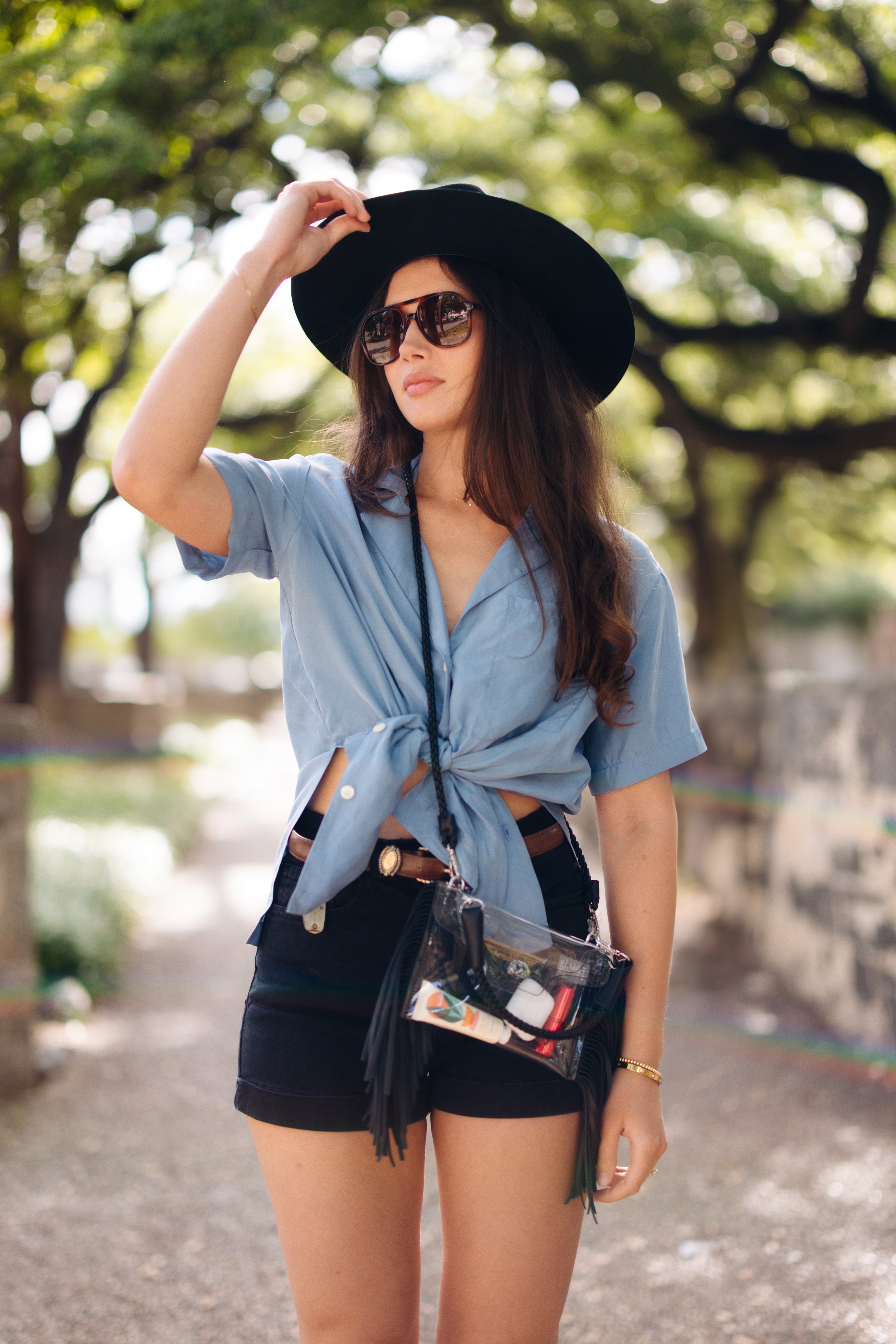 Woman wearing a black hat, sunglasses, and a blue top with a black skirt in an outdoor setting. With the clear bag adorning her as an accessory.