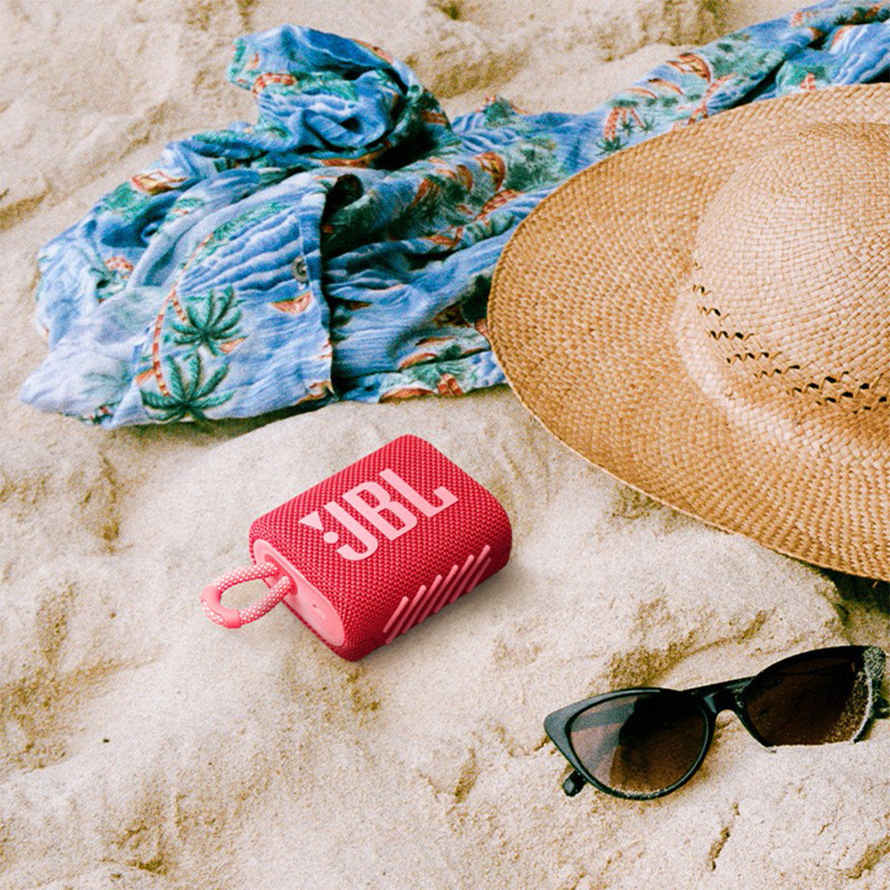 Red JBL Go 3 speaker on beach sand. Rugged IP67 waterproof design for beach and travel use.